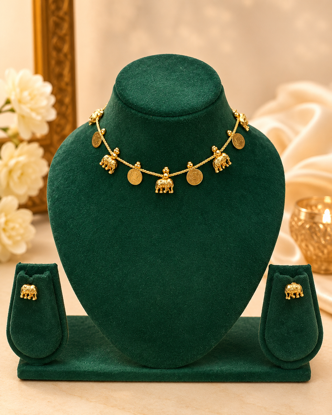 Gold jewelry set displayed on a dark green velvet stand, including a necklace with dangling elephant motifs, matching stud earrings, and a small box, against a softly lit beige background with decorative flowers and fabric.