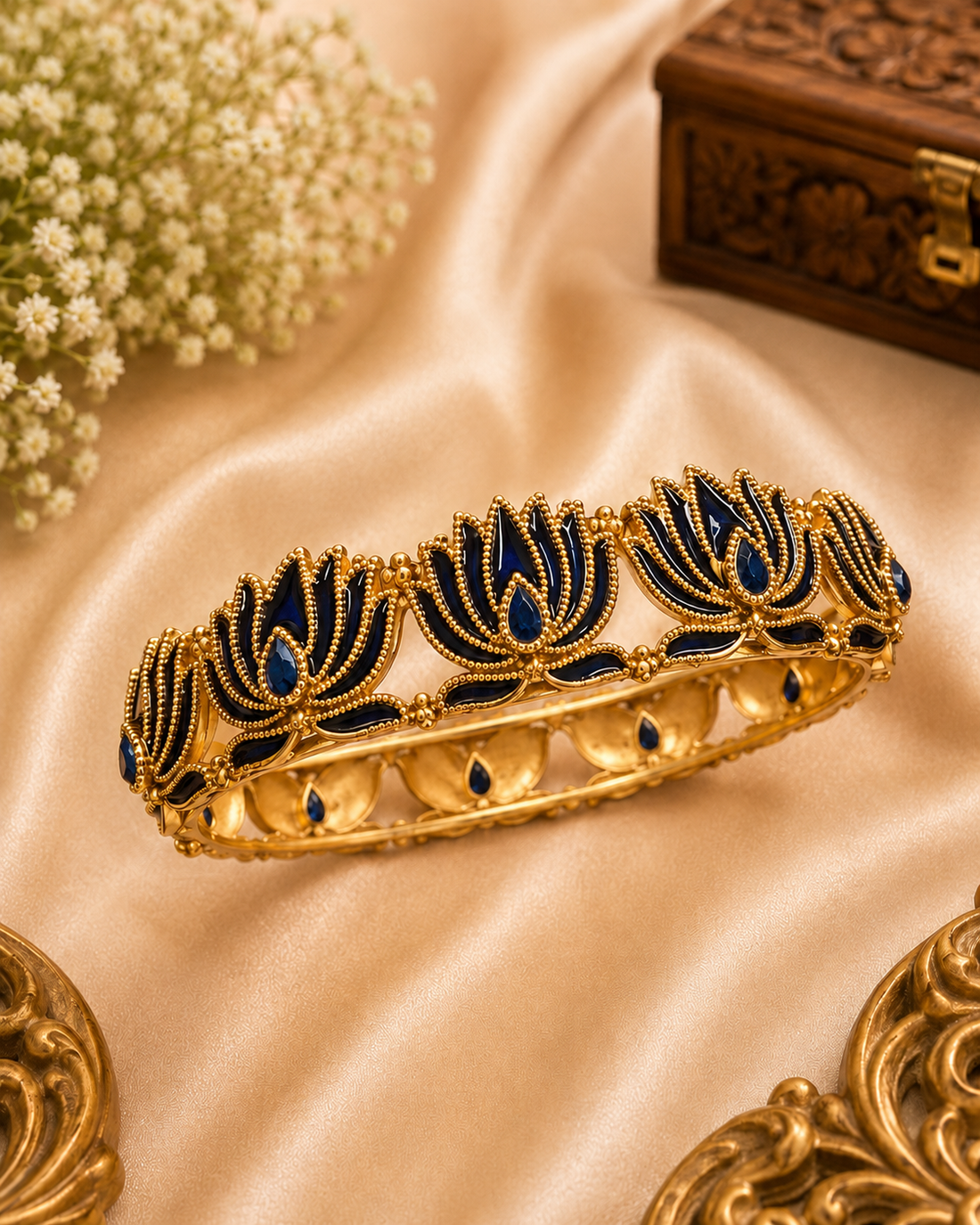 A gold-plated traditional Indian bangle bracelet decorated with blue enamel lotus motifs, displayed on a draped beige satin fabric with decorative floral and wooden props.