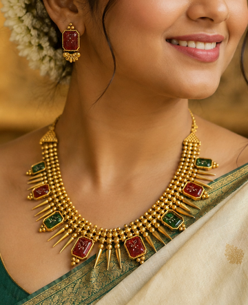 A woman wearing a gold-toned traditional necklace with red and green stone accents, matching earrings, and a cream saree with a green border, photographed in an ornate indoor setting.