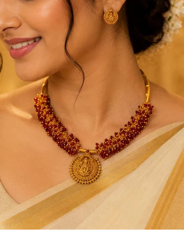 A close-up of a woman wearing a gold and red beaded necklace with a round pendant, matching gold earrings, and a cream-colored sari against a warm golden background.