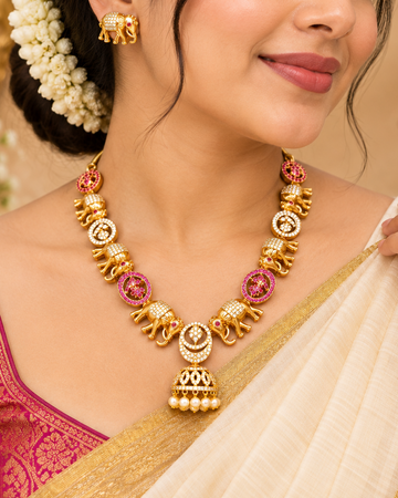 A woman wearing a gold-toned traditional necklace with elephant and floral motifs, matching gold earrings, and a cream saree with a magenta blouse.