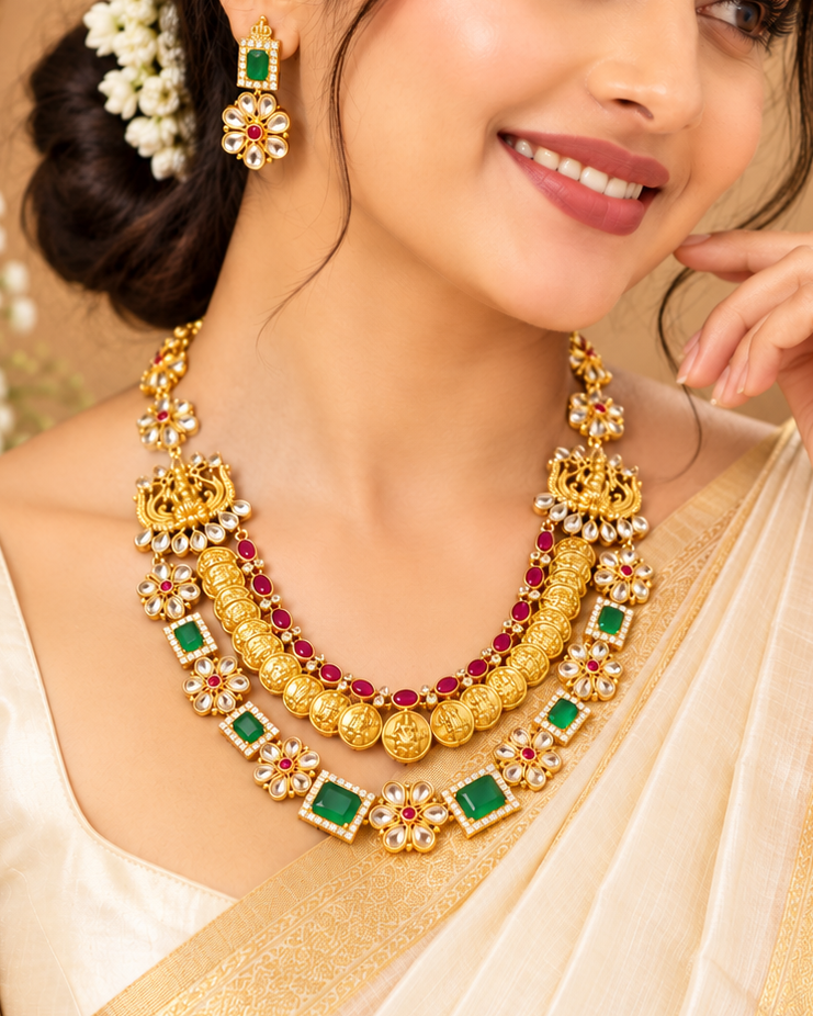 A woman wearing an ornate traditional gold necklace with green and red stones, matching gold earrings, and a cream saree with a gold border, posed against a warm beige background with decorative floral elements.