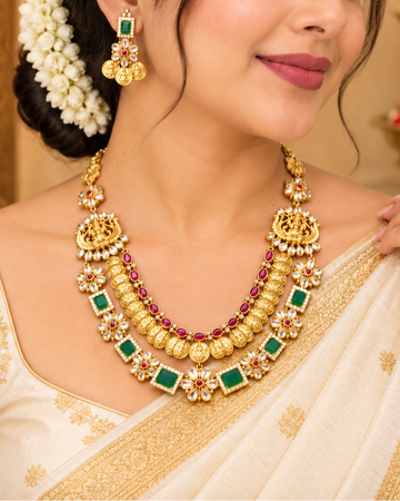 A woman wearing an ornate gold-toned traditional necklace with green stones and red gem accents, matching drop earrings, and a cream saree with gold embroidery in a warm indoor setting.