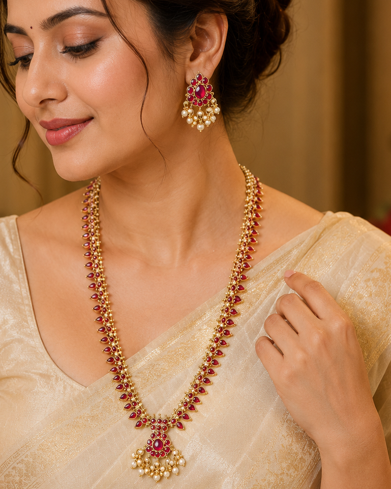 A woman wearing a traditional Indian outfit in beige and gold, styled with a matching gold and red beaded necklace and matching red and gold earrings.