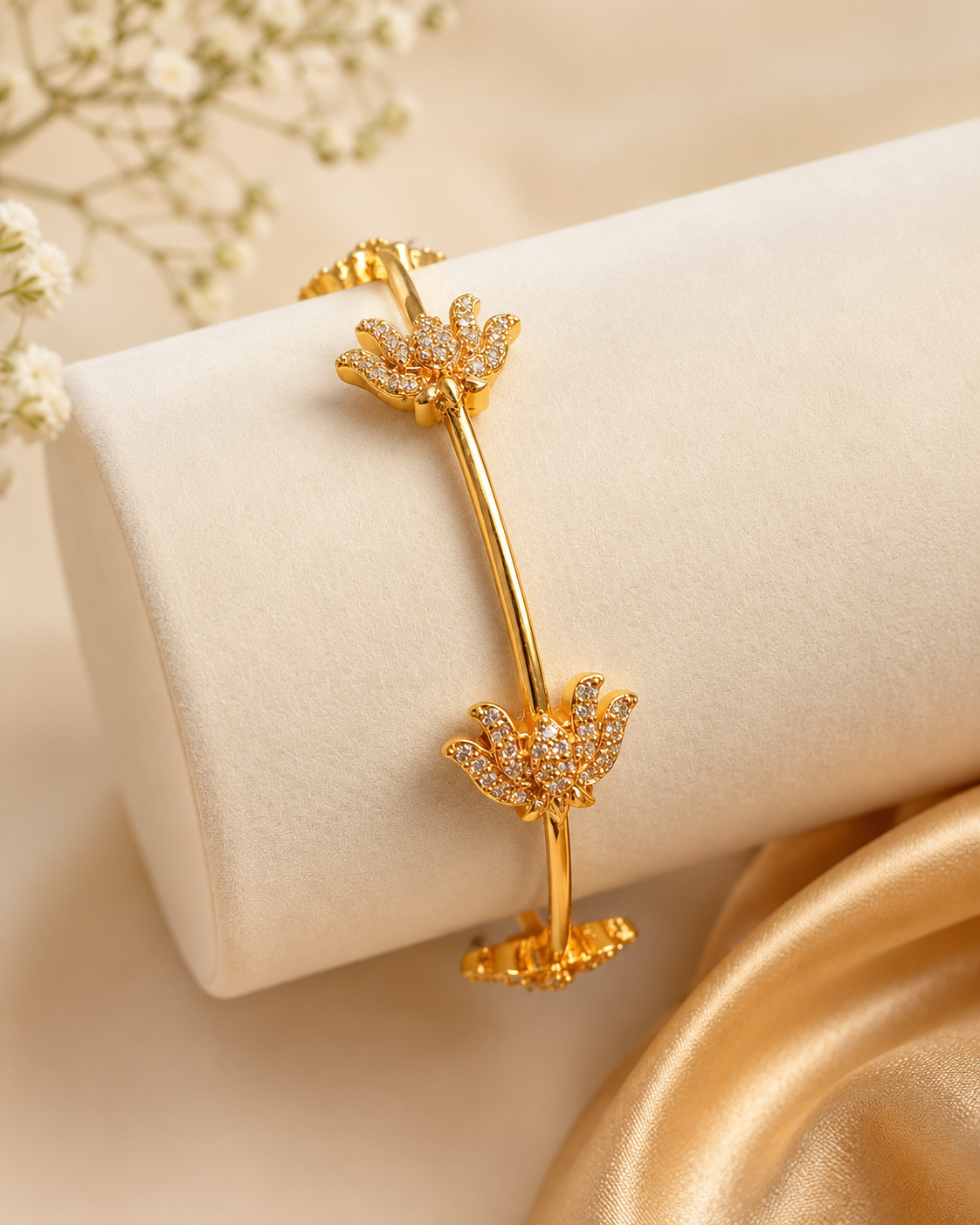 A gold bangle bracelet with two floral motifs paved with small clear stones, displayed on a cream jewelry stand with soft beige fabric and white flowers in the background.