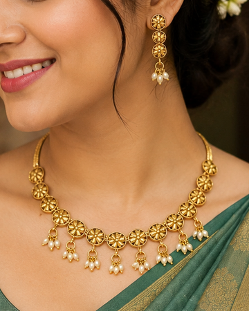 A close-up of a woman wearing a gold-toned traditional necklace and matching drop earrings with small white pearl-like accents, paired with a green sari.