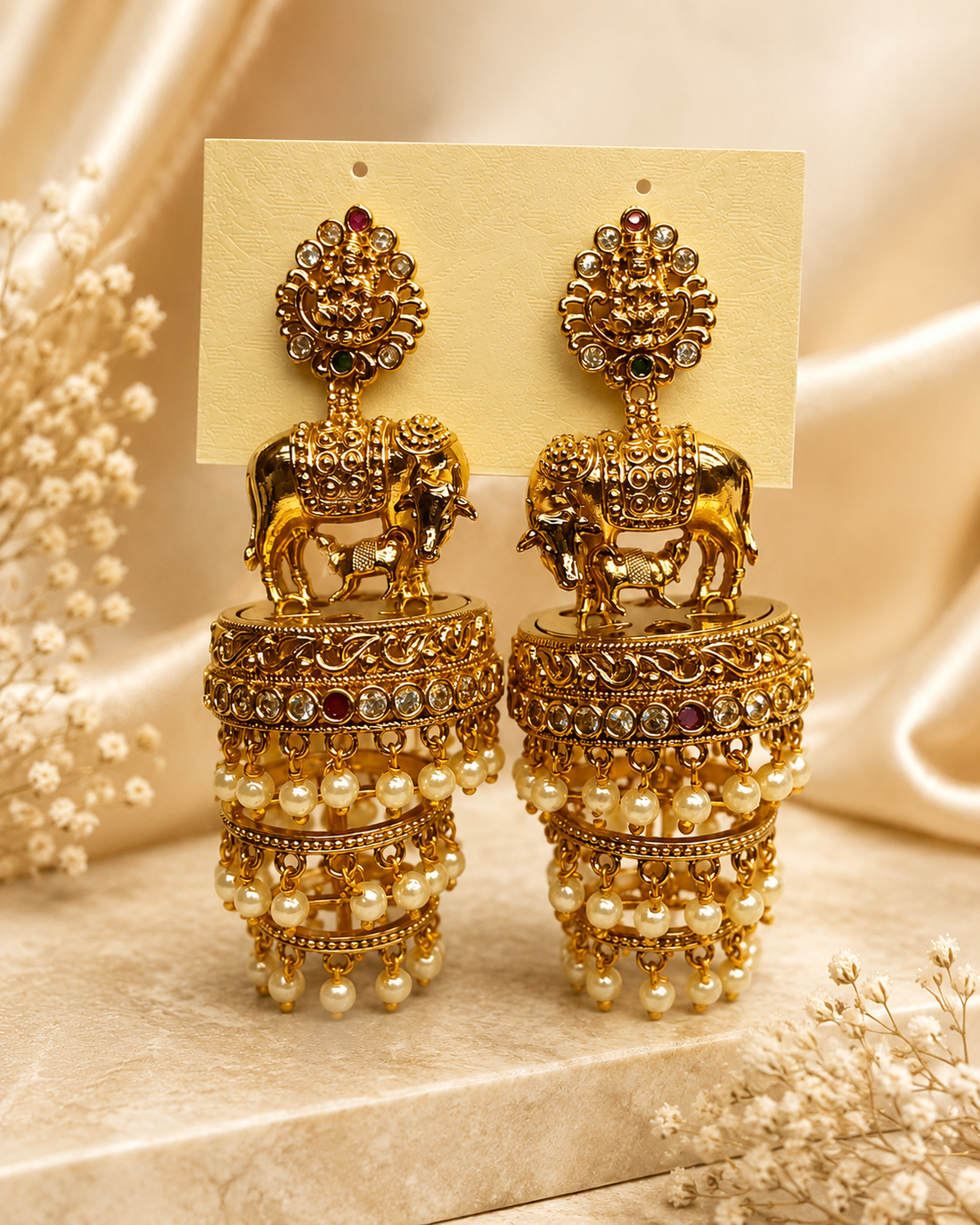 A woman wearing ornate gold-toned traditional earrings with dangling pearl-like beads and decorative motifs, paired with a light beige sari blouse in a softly lit indoor setting.