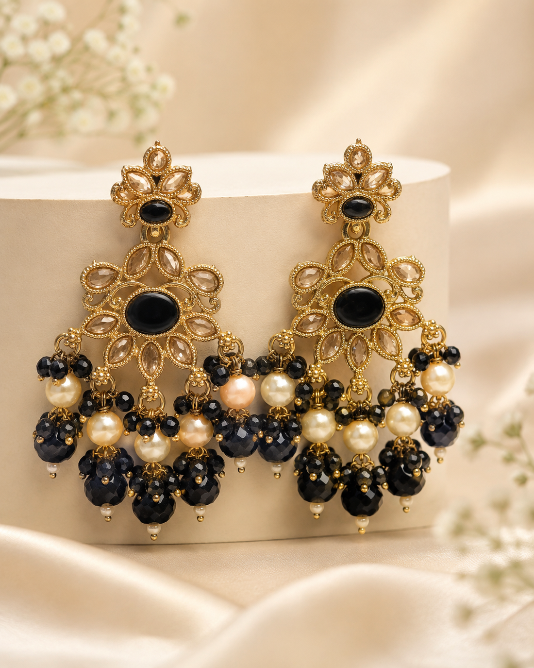 A woman in profile wearing ornate gold-toned traditional earrings with black and white bead drops, paired with a cream-colored embroidered saree blouse.