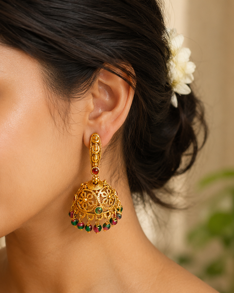 A close-up lifestyle shot of a woman wearing an ornate gold Jhumka earring adorned with red and green stones, paired with a cream and gold saree.