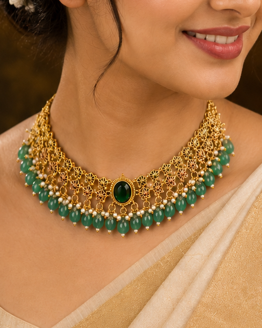 A woman wearing a traditional Indian outfit with a beige saree and a gold-toned statement necklace adorned with green beads and a central green stone, set against a warm, dark background with decorative elements.