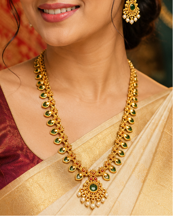 A woman wearing a traditional Indian saree in beige and maroon with gold jewelry, including a gold necklace with green stone accents and matching earrings, posed against a background of ornate jewelry displays.