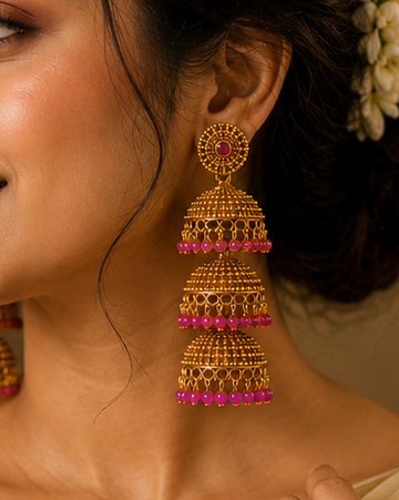 A woman wearing ornate gold jhumka earrings with pink and red beaded tassels, styled with a cream and gold saree and a dark green blouse.