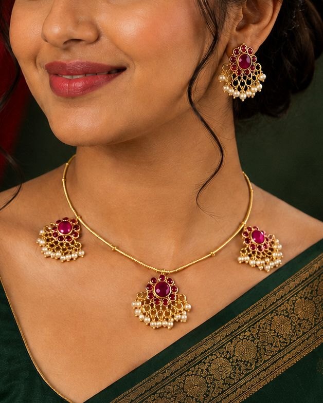A woman wearing a dark green saree with a gold border and matching gold jewelry featuring red and white stones, posed against a dark green background with red drapery.