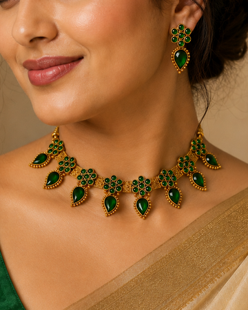 A woman wearing a gold and green jewelry set with a matching necklace and earrings, paired with a green blouse and a beige saree, set against a warm beige background.