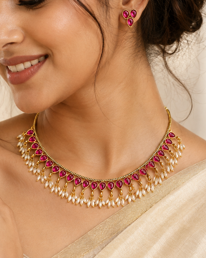 A woman wearing a gold-toned traditional necklace with red stones and white pearl-like drops, paired with matching red and gold earrings, in a warm neutral indoor setting.
