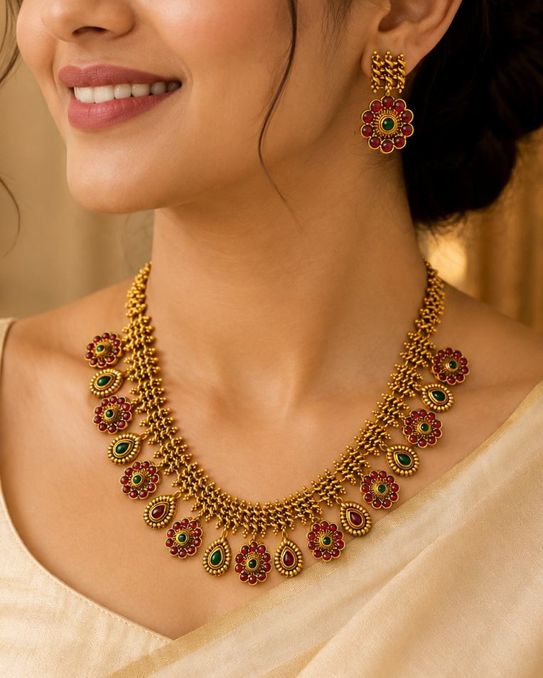 A woman wearing an ornate gold-toned traditional necklace with red and green stone accents and matching drop earrings, shown in a warm indoor setting.