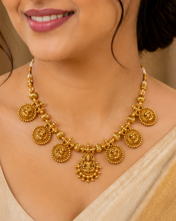 A woman wearing a traditional gold necklace with circular motifs and a matching beige saree, shown in a warm, elegant indoor setting.