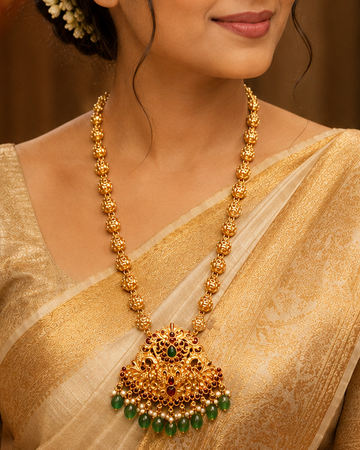 A woman wearing a traditional Indian saree in beige and gold, styled with a large gold necklace featuring a prominent gold and green pendant, set against a warm, ornate background.