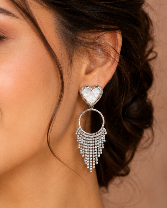 A close-up of a woman wearing a pair of silver-toned statement earrings with a heart-shaped crystal top and dangling crystal fringe, against a warm beige background.