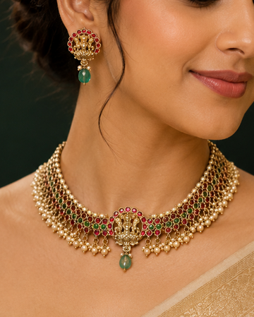 A woman wearing an ornate gold-toned traditional necklace with green and red stone accents, matching earrings, and a beige sari against a dark background with red drapery.