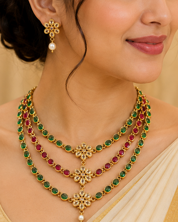 A woman wearing a gold-tone layered necklace with green and red stones, matching earrings, and a cream saree with a gold blouse, posed in an elegant indoor setting.