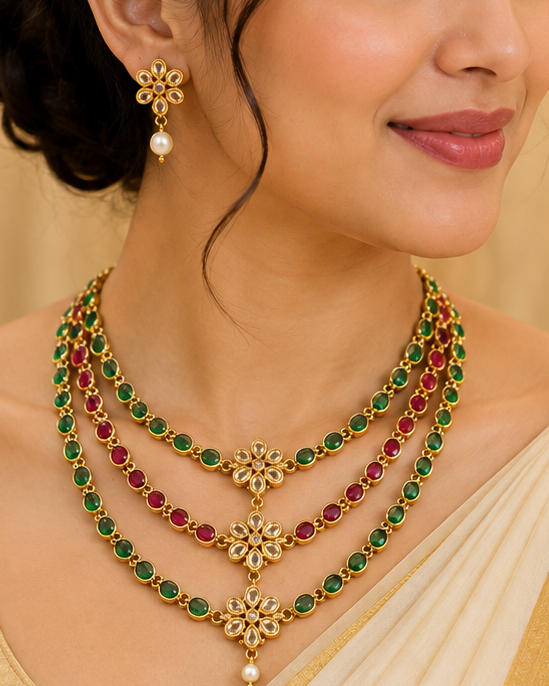 A woman wearing a gold-tone layered necklace with green and red stones, matching earrings, and a cream saree with a gold blouse, posed in an elegant indoor setting.