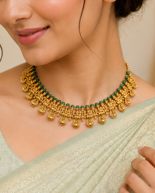 A woman wearing a light sage green saree and matching blouse, styled with a gold-toned traditional necklace and bracelet in an elegant indoor setting.