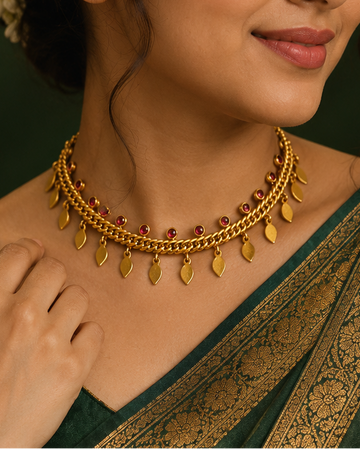 A woman wearing a dark green sari with gold zari embroidery and a matching gold leaf necklace, posed against a dark green background with warm golden lighting.