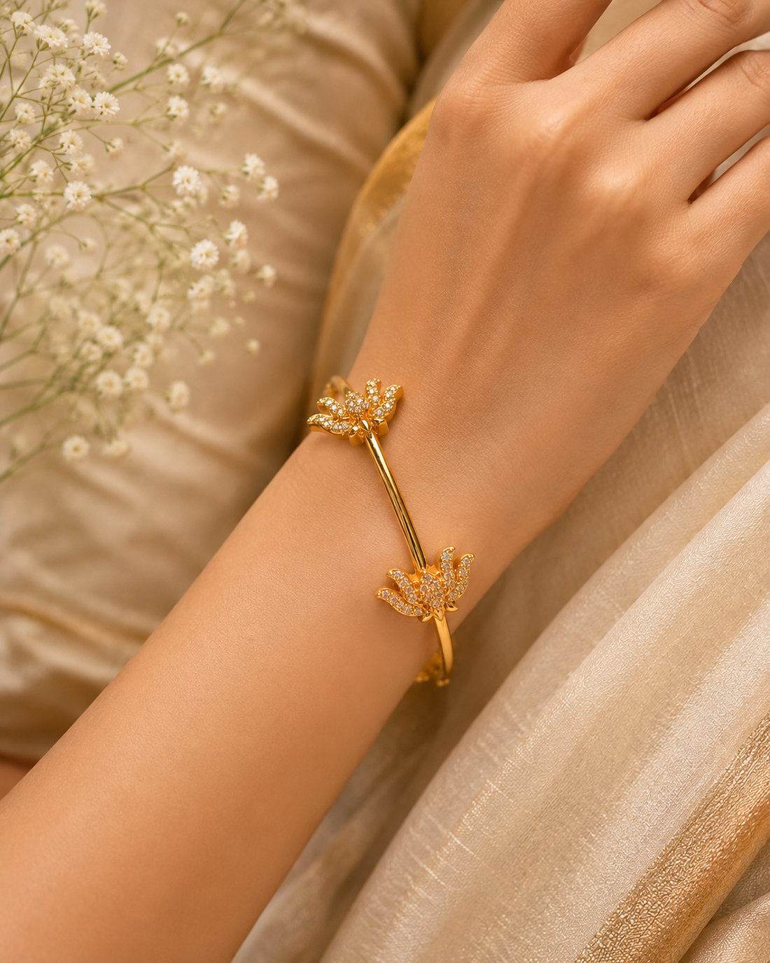 A gold-toned wrap bracelet with two floral motifs set with small clear stones, worn on a person's wrist against a soft beige fabric background with baby's breath flowers.