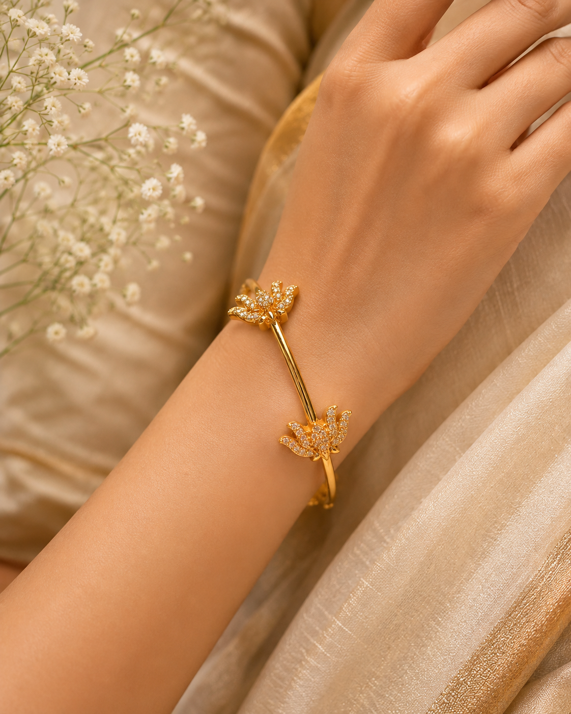A gold-toned wrap bracelet with two floral motifs set with small clear stones, worn on a person's wrist against a soft beige fabric background with baby's breath flowers.