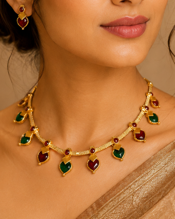 A woman wearing a gold-toned necklace with alternating red and green heart-shaped stones, paired with matching earrings and a gold saree, set against a warm, elegant background.