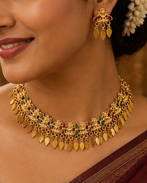 A woman wearing a deep maroon sari with gold zari embroidery, paired with a matching gold necklace and earrings, set against a warm, traditional background.