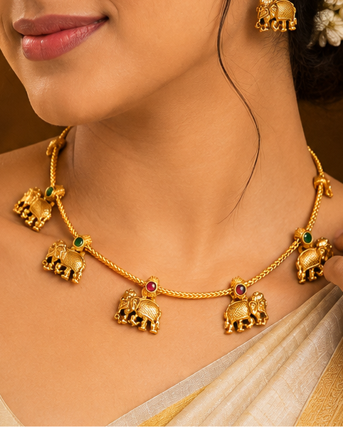 A woman wearing a gold elephant necklace and matching earrings, styled with a cream and gold saree, set against a warm, traditional background.