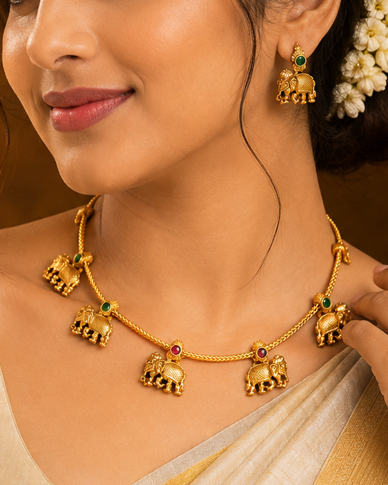 A woman wearing a gold elephant necklace and matching earrings, styled with a cream and gold saree, set against a warm, traditional background.