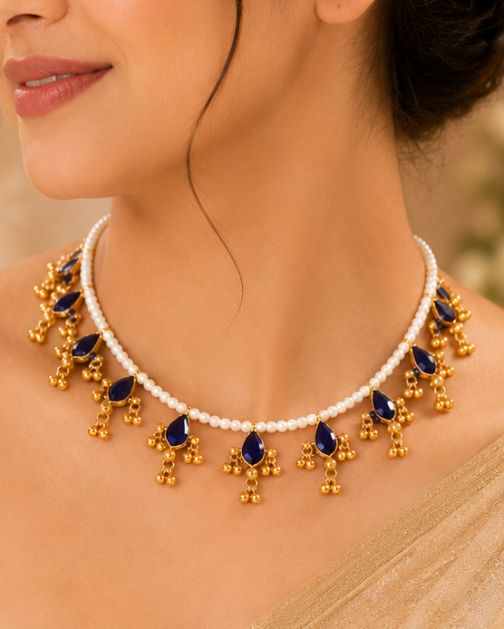 A woman wearing a gold and navy blue beaded necklace with white pearl-like accents, styled with a beige saree in a warm, elegant indoor setting.
