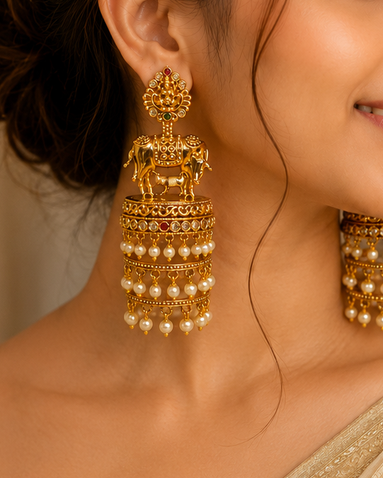 A woman wearing ornate gold-toned traditional earrings with dangling pearl-like beads and decorative motifs, paired with a light beige sari blouse in a softly lit indoor setting.