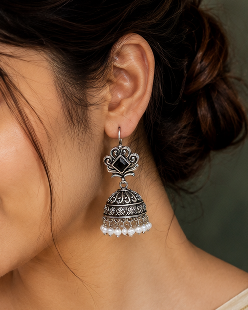 A woman wearing a cream-colored saree and matching blouse, shown in a close-up portrait. She is wearing a traditional silver-toned jhumka earring with black stone detailing and white pearl accents.