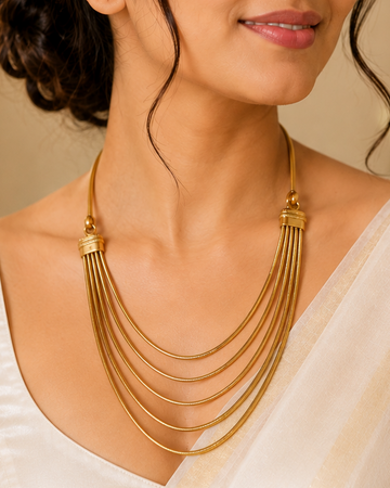 A woman wearing a multi-strand gold necklace with a layered, tiered design over a white sari, shown in a warm, elegant indoor setting.