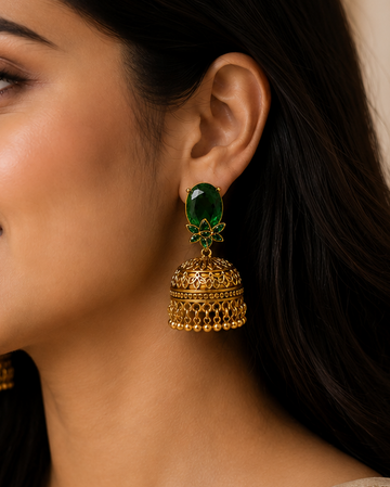 A woman in a beige sari wearing ornate gold jhumka earrings with green stone accents, shown in a close-up portrait against a soft beige background.
