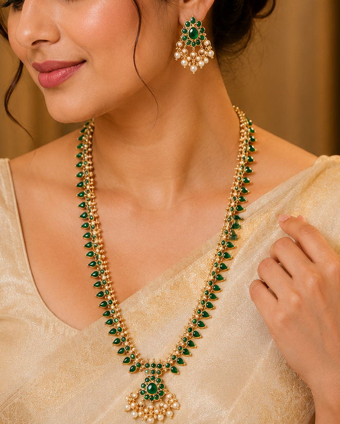 A woman wearing a traditional Indian outfit with a beige saree and blouse, styled with a green and gold beaded necklace and matching green and gold earrings.