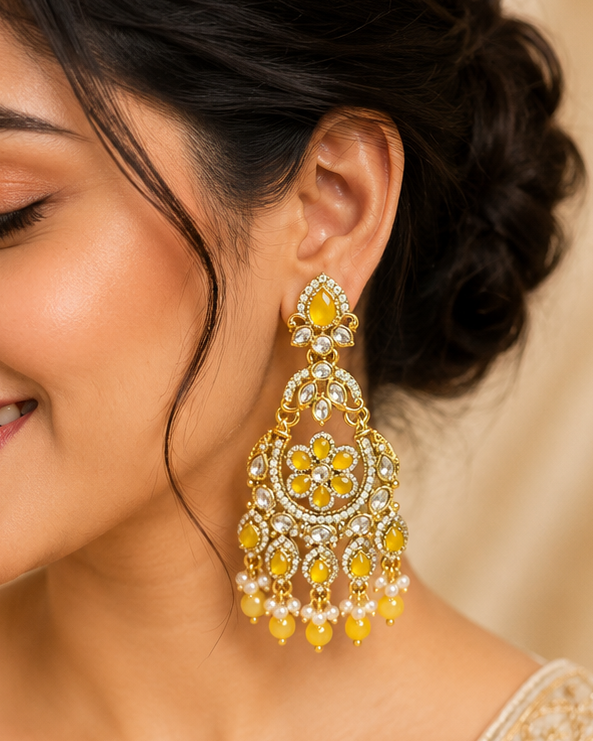 A woman wearing a large, ornate gold and yellow chandelier earring with intricate floral details and hanging pearl-like drops, paired with a cream-colored embroidered traditional top against a soft beige background.