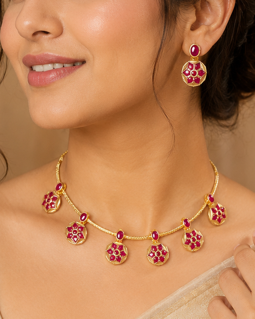 A woman wearing a gold-toned necklace and matching earrings with pink stone accents, styled with a light beige saree against a warm beige indoor backdrop.