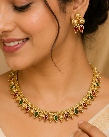 A woman wearing a gold-toned traditional necklace with green and red stones, matching earrings, and a cream-colored saree, shown in a warm indoor setting.