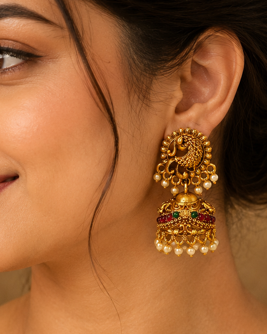 A woman in a beige saree wearing a large, ornate gold-toned traditional Indian jhumka earring with green and red stone accents, shown in a close-up portrait.