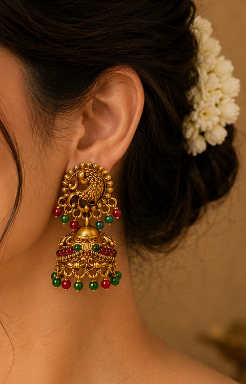A woman in a beige saree wearing a large, ornate gold-toned traditional Indian jhumka earring with green and red stone accents, shown in a close-up portrait.