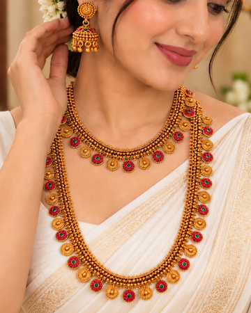 A woman wearing a traditional Indian gold necklace with red stone accents and matching gold earrings, styled with a white saree in a warm, decorated indoor setting.