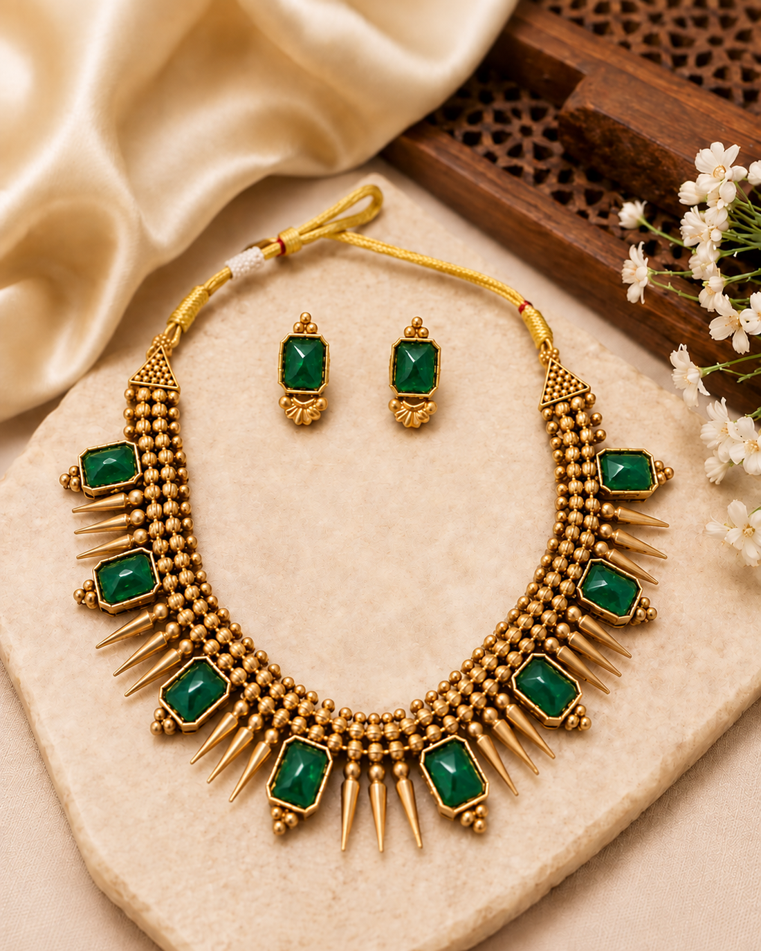 A woman wearing a gold-toned traditional necklace with red and green stone accents, matching earrings, and a cream saree with a green border, photographed in an ornate indoor setting.