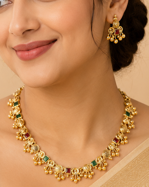 A woman wearing a gold-toned traditional necklace and matching earrings, styled with a beige saree against a warm beige background.