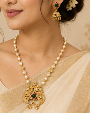 A woman wearing a traditional Indian outfit in beige and gold, featuring a pearl necklace with a gold pendant and matching gold earrings, set against a warm, softly blurred background.