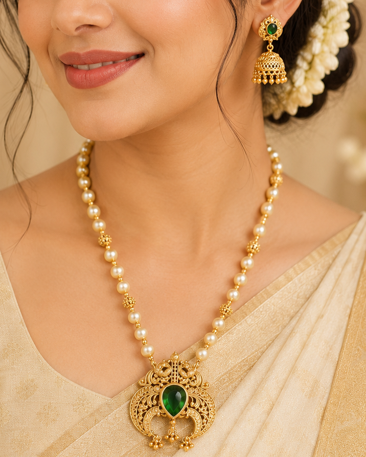 A woman wearing a traditional Indian ensemble, featuring a gold and green pendant necklace, matching gold and green jhumka earrings, and a cream-colored sari.